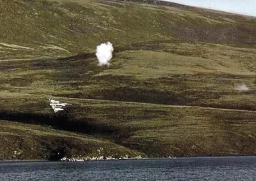 Fotografía del Avión Dagger de la Fuerza Aérea Argentina maniobrando dentro del estrecho de San Carlos para atacar a los buques ingleses mientras se ven explosiones del fuego antiaéreo.