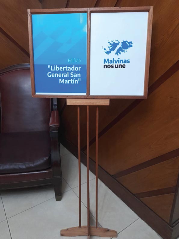 La fotografía muestra un atril con dos carteles, el primero con la leyenda del nombre del edificio: “Libertador General San Martín” y el segundo que dice: “Malvinas nos une” cercano al ascensor para los oficiales de rango superior.