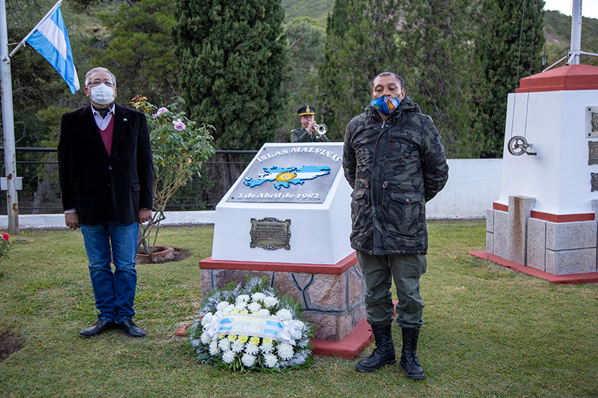 La fotografía muestra a dos Veteranos de la Guerra de Malvinas parados al lado del monumentos a los caídos en la Guerra de Malvinas con la ofrenda floral