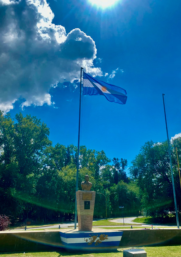 La fotografía muestra el monumento a los veteranos y caídos en Malvinas en Cosquín, que remata en la parte superior con el busto del Capitán Aviador Omar Jesús Castillo y detrás un mástil con la bandera argentina flameando.